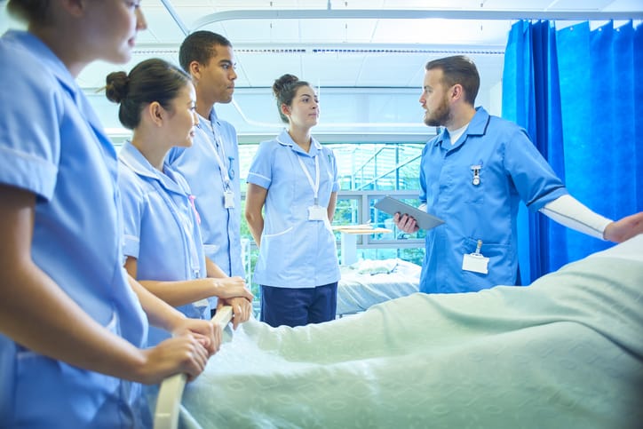 A senior male staff nurse demonstrates the medical mannequin to a group of medical student nurses . They are all standing around the hospital bed listening to him .